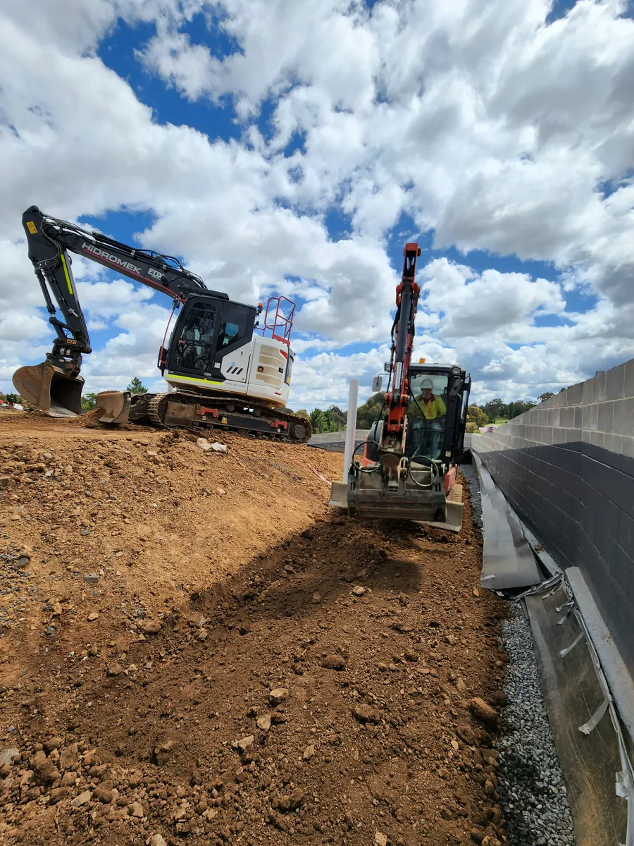 Excavator and skid steer shaping a batter beside a retaining wall